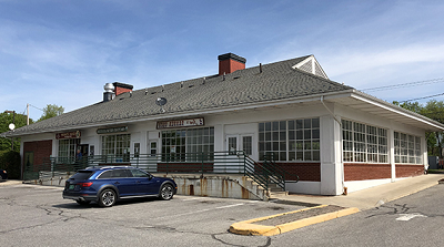 Single-story brick building with white munton windows, green railing up stairs and ramp, edge of paved parking lot.