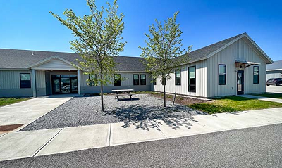 Single-story office building with ell addition, two entrances, with concrete sidewalks, young trees and picnic table out front. 